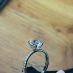 Image of a hand holding a diamond oval engagement ring above a wooden table.